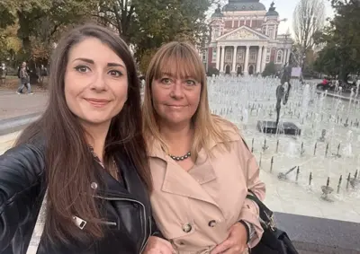Two women smile for a selfie in front of a fountain with a neoclassical building in the background; one with dark hair in a black leather jacket, the other with blonde hair in a beige trench coat and necklace.