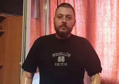 Bearded man in a black T-shirt reading Medellín 85 Critic, standing indoors with red curtains behind him.